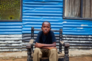 An African boy sitting in a broken chair with his hands on his knees while looking seriously at the camera.