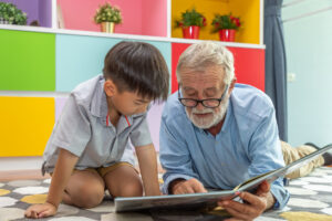 Happy boy grandson reading book with old senior man grandfather at home