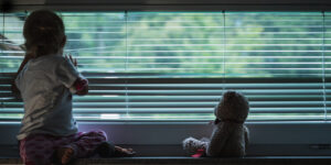 Toddler girl looking out the window through lowered venetian blinds with her teddy bear sitting next to her