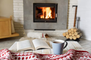 Hygge concept with open book and cup of tea near burning fireplace