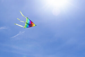 The multi-colored strips of a rainbow kite flies in the clear blue sky in the rays of the sun.