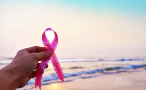 Breast cancer awareness month concept, hands holding pink ribbon against beach with blue sky. Blur defocused background, copy space text area.