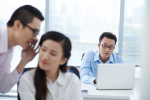 Unhappy young businessman looking at gossiping colleagues