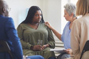 Portrait of sad African-American woman listening to psychologist during support group meeting with people siting in circle and comforting her