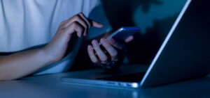 close up young man hand holding smartphone for access by unlock on screen and using laptop for register website on internet in the late night , internet addiction concept