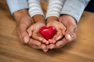 Family holding small red heart in hands on wooden background. Top view of father and daughter hands protect heart. High angle view of indian man and little girl hands holding red heart: adoption foster family, hope, gratitude and insurance concept