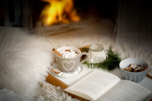 Cup of cocoa, hot chocolate with marshmallows on a tray with book, lantern and fir branch on a sofa or bed with blankets