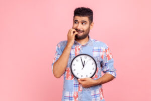 Too late! Portrait of impatient anxious man with beard in blue shirt holding big clock and biting nails, nervous about delayed meeting or deadline. Indoor studio shot isolated on pink background.