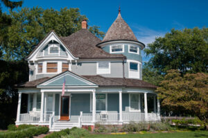 Beautiful gray traditional victorian house. House has an American Flag haning over the porch and shows a beautiful garden with flowers and trees. Set against a cloudless blue sky