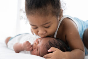 Cute African American little girl kissing on newborn baby cheek on white bed at home. Little girl takes care of infant baby with kindly