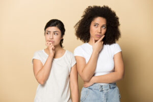 Puzzled thoughtful diverse girls touching chin with fingers, planning future, two female customers pondering offer, making decision, looking aside, standing isolated on brown background