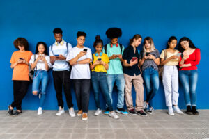 Group of multiracial teenager high school students stand leaning against blue wall looking at cellphone. Social media addiction concept. Education concept.