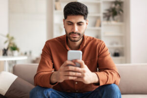 Portrait of young Middle Eastern man sitting on couch with smartphone, chatting or checking email, home interior, free copy space. Focused guy using brand new mobile phone, checking social media