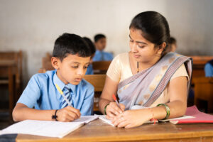 Teacher in classroom helping kid for studying at desk - concept of personal care, mentorship and coaching.