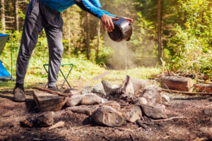 Man extinguishing campfire with water from cauldron in summer forest. Put out campfire by tent. Traveling fire safety rules