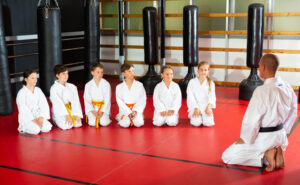 Group of karate kids in white kimono kneeling in front of their trainer on gym floor.