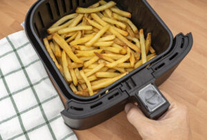 HAND HOLDING AIR FRYER BASKET WITH FRESHLY MADE FRENCH FRIES OR FRIED POTATOES IN THE KITCHEN COUNTERTOP. TOP VIEW.