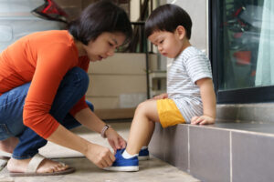 Asian Chinese mother helping her son put shoes on at home outdoors. First day of school.