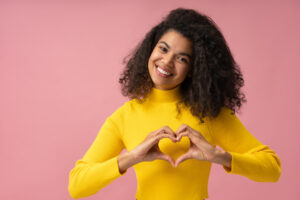 Portrait of beautiful smiling African American woman with toothy smile showing heart shape near face isolated on pink background