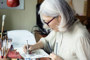 Asian senior woman artist sketching. Focus on face, blurred workspace in background. Art background, text space
