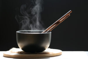 Selective focus of smoke rising with hot soup of instant noodles in cup and chopsticks on dark background