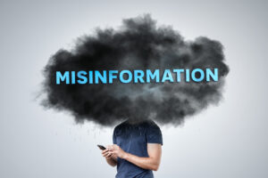 A person stands holding a smartphone, with their head obscured by a dark cloud labeled "Misinformation." The image symbolizes the concept of fake news and digital deception, illustrating the impact of misinformation in the digital age.