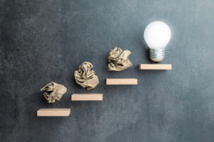 Conceptual image of crumpled paper balls as steps leading to a light bulb, symbolizing failure as part of the creative or problem-solving journey toward success