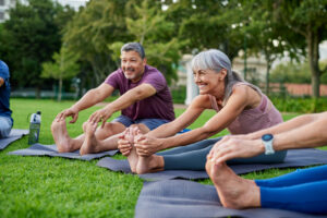 Seniors doing stretching exercises together in circle on yoga mats at city park. Elderly people smiling while bending forward to touch toes. Group of women and men during yoga session for fitness in the park.